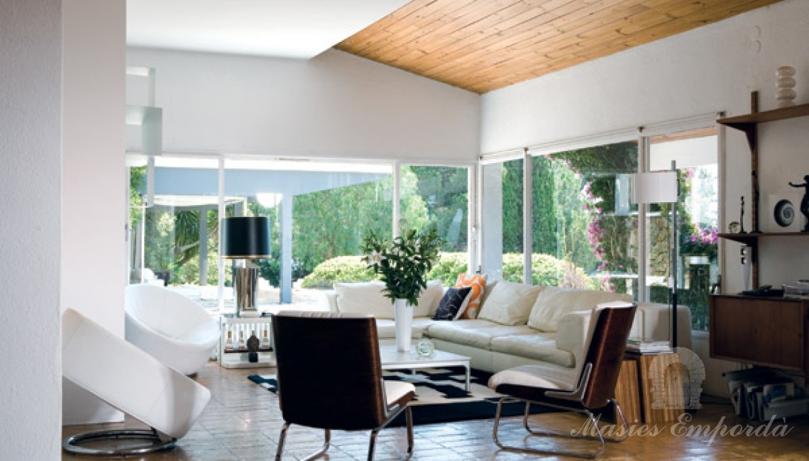 Prospectiva del salón de estar de la casa con acristalamiento y vistas al jardín con detalles de la cubierta en madera clara que resalta del color blanco del resto de techos