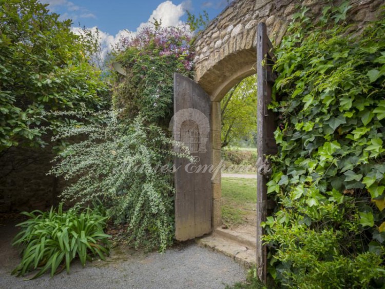 Portón de madera de entrada al partió de la masía con el jardín en flor aun lado del portón y una exuberante enredadera en el otro 