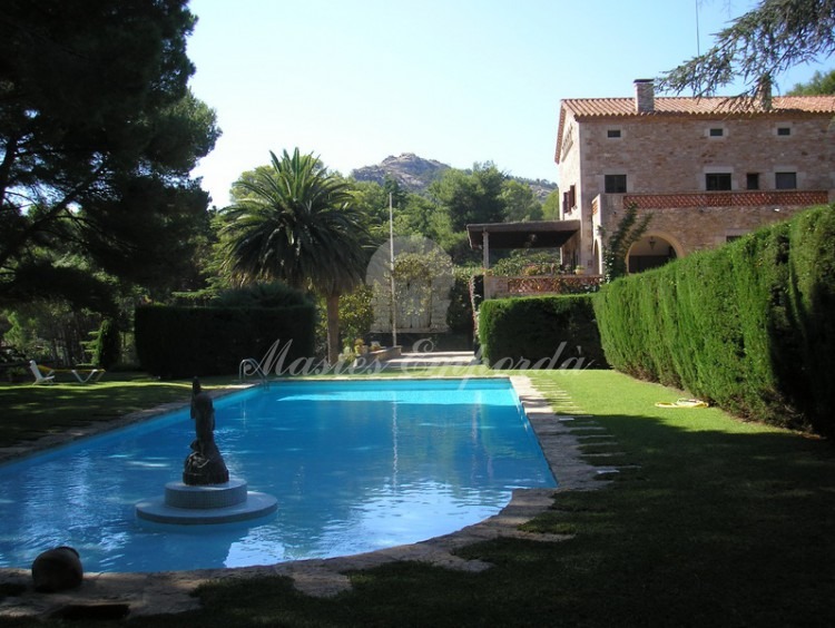 Vista de la piscina con escultura en piedra caliza en su interior y de fondo la casa y los jardines de la casa