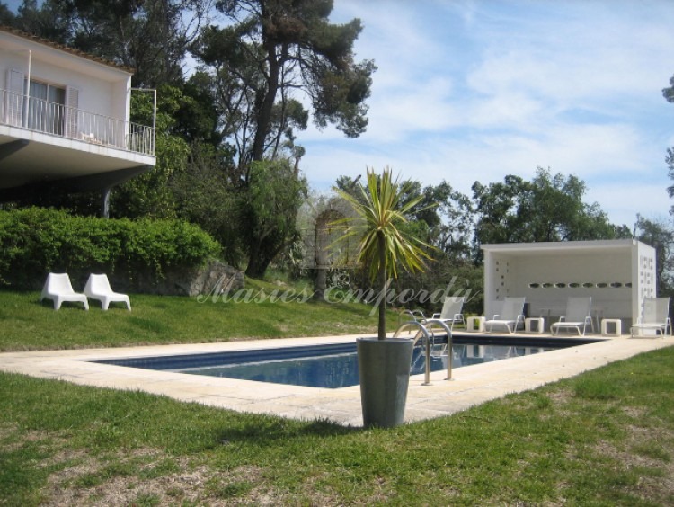 Detalle de la zona de la piscina con pequeño porche en blanco que contrata con el verde del maravilloso jardín de la propiedad